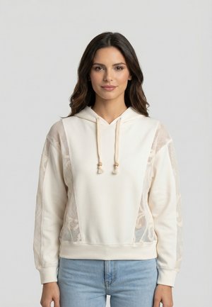 Jonge vrouw met lang donker haar die een crème hoodie met mesh-details en lichtblauwe jeans draagt, staand tegen een effen lichte achtergrond.