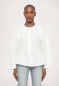 Blusa de algodão branca com decote redondo, mangas bufantes longas e pregas verticais. Combinada com jeans de ganga clara.