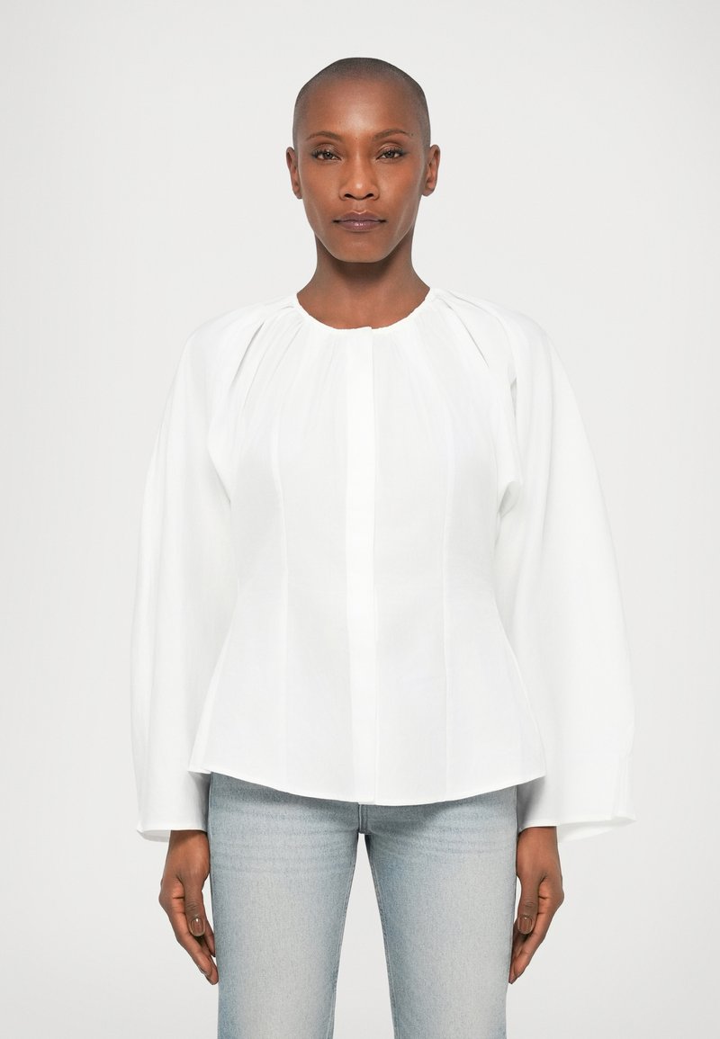 Blusa de algodão branca com decote redondo, mangas bufantes longas e pregas verticais. Combinada com jeans de ganga clara.