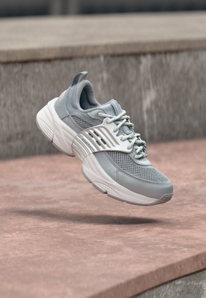 Lichtgrijze sportieve sneaker met witte zool en banddetail, hangend boven een rozeachtige betonnen ondergrond met een grijze stenen achtergrond.