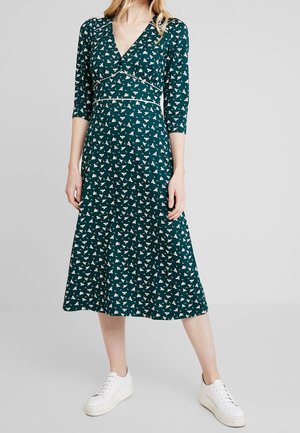 Femme portant une robe bleu sarcelle mi-longue à motifs d'oiseaux blancs et manches trois-quarts, associée à des baskets blanches à plateforme.