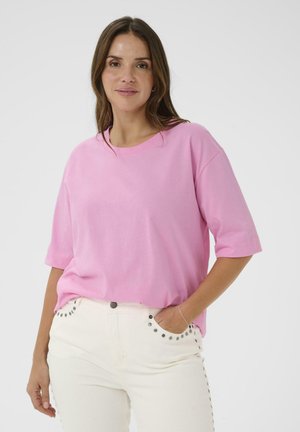 Kvinde iført en løs, lyserød kortærmet skjorte og hvide bukser med metalnitte detaljer, stående med den ene hånd i lommen mod en ensfarvet baggrund.