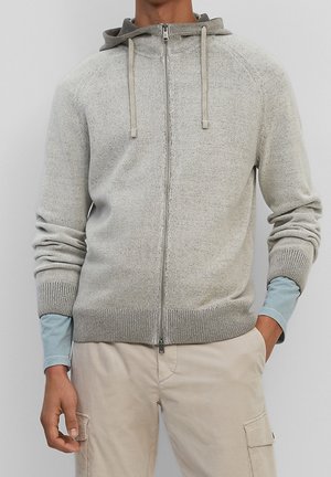 Haut de sweat à capuche zippé gris clair avec une capuche bicolore, en tissu tricoté texturé, avec des poignets côtelés et une coupe droite. Il est équipé de cordons de serrage pour un ajustement.