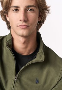 Giacca zip-up verde oliva con una leggera trama a costine e un logo ricamato nero sul petto. Indossata sopra una t-shirt nera.