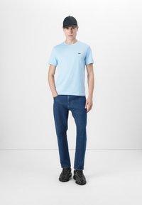 T-shirt bleu clair à manches courtes avec un col rond, associé à un jean bleu foncé et des baskets noires. Casquette noire portée sur la tête.