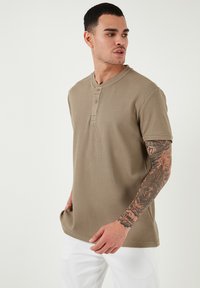 Khaki színű, rövid ujjas henley póló texturált anyagból, három gombbal a nyaknál, laza szabással és egyenes aljjal.