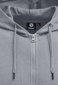 Grauer Reißverschluss-Hoodie aus glattem Stoff. Ausgestattet mit einem silbernen Reißverschluss, verstellbarem Kordelzug an der Kapuze und einem Etikett mit Markenlogo und Größeninformation.