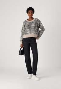Pull en tricot beige et marine avec un motif géométrique, associé à un jean noir et des baskets blanches. Le mannequin tient un petit sac noir.
