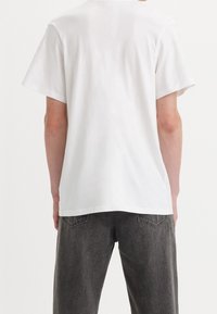 Biały bawełniany T-shirt z krótkimi rękawami i okrągłym dekoltem, noszony z ciemnoszarymi dżinsami, prezentujący widok z tyłu oraz minimalistyczny design.