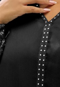 Blouse noire avec un tissu texturé, un design en V et une rangée de petites clous en argent le long de la couture avant. Détail de manches en sequins visible.