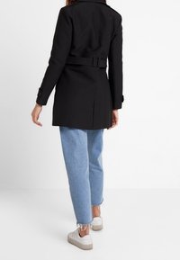 Osoba oblečená v černém kabátu s páskem, světle modrých džínech s roztrhanými lemy a bílých teniskách, stojící na jednobarevném bílém pozadí.