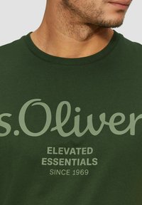 Man wearing dark green t-shirt with "s.Oliver ELEVATED ESSENTIALS SINCE 1969" printed on front, cropped at neck and chin.
