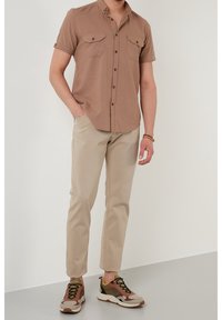 Mann, der ein braunes kurzärmliges Hemd mit Knöpfen, beige Hose und mehrfarbige Sneaker trägt und vor einer schlichten weißen Wand steht.