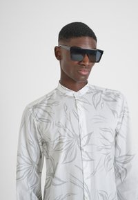 Camisa ligera de manga larga con fondo blanco y patrones de hojas grises, combinada con unas grandes gafas de sol negras.