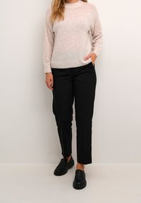 Frau in einem hellbeigen Pullover, schwarzen geraden Hosen, schwarzen Slippern, steht mit einer Hand in der Tasche vor einem schlichten Hintergrund.