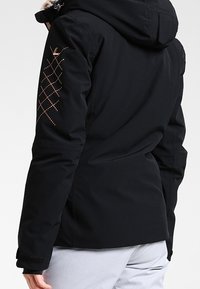 Schwarze isolierte Jacke mit Kapuze, ausgestattet mit rautenförmigen Akzenten in Roségold am linken Ärmel und verstellbaren Bündchen.