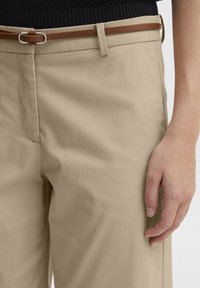 Beige bomullsbukser med slim fit. Har et brunt belte med en sølvspenne og en glatt tekstur, som viser subtile folder.