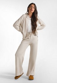 Beige loungewear set bestaande uit een ritssluiting hoodie en wijde broek. Zacht, textuurstof met subtiele glinstering accenten. Bruine slippers.