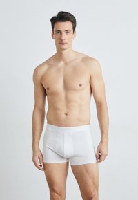 Bílé bavlněné boxerky s hladkým povrchem, s logem potištěným na pase a přiléhavým střihem, modelované mužem v neutrálním prostředí.