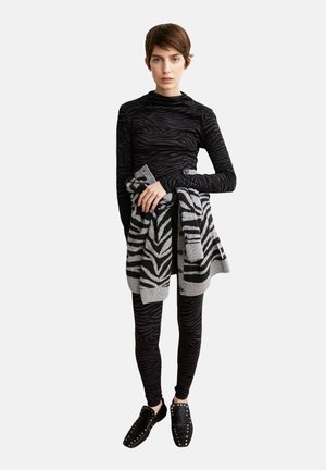 Schwarzes Langarmoberteil mit einem dezenten Zebramuster, kombiniert mit schwarzen Leggings. Graue Strickjacke mit einem auffälligen Zebramuster, um die Taille gewickelt.