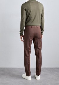 Braune maßgeschneiderte Hosen aus glattem Stoff, kombiniert mit einem schlanken, olivgrünen Langarmshirt und weißen Sneakern.