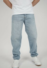 Person, der hellblaue, locker sitzende Jeans und weiße Sneaker trägt und vor einem schlichten Hintergrund steht.