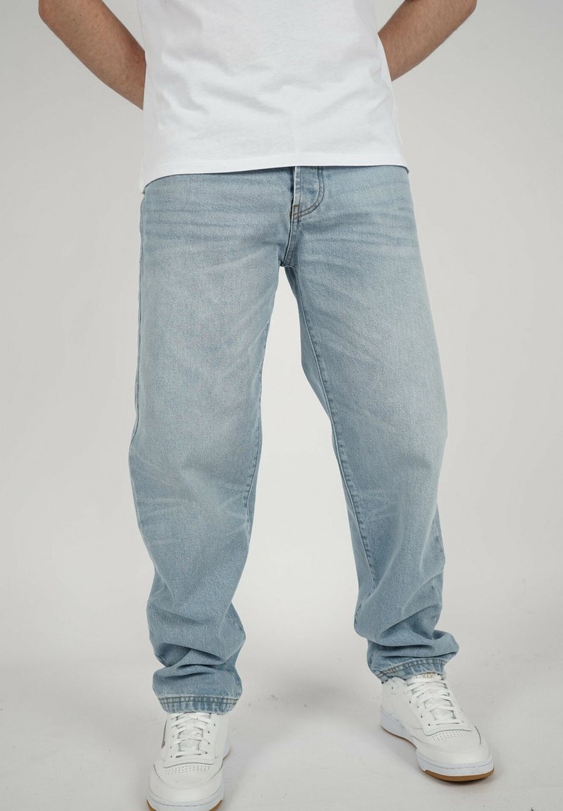 Person, der hellblaue, locker sitzende Jeans und weiße Sneaker trägt und vor einem schlichten Hintergrund steht.