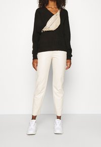 Schwarzer, gestrickter V-Ausschnitt-Pullover, kombiniert mit weißen, schmalen Hosen und weißen Sneakers. Eine beigere Umhängetasche mit Reißverschlüssen wird über die Brust getragen.