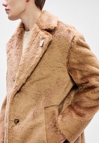 Kafijas krāsas mākslīgās kažokādas mēteli ar iegrieztiem atloka, lieliem pogām un sānu kabatām. Tekstūra ir mīksta ar vieglām krāsu variācijām.