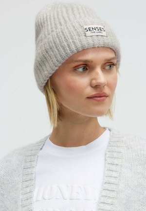 Hellgraue, gestrickte Mütze mit gerippter Struktur und einem rechteckigen Aufnäher mit der Aufschrift "SENSES THE LABEL." Weiches Wollmaterial. Eng anliegendes Design.