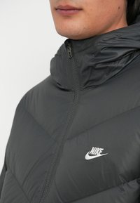Tamno siva jakna od puf materijala s visokim vratom, zatvaranjem na zip, i bijelim Nike logotipom. Ima kviltanu teksturu i lagani materijal.