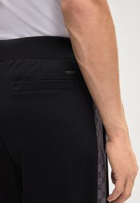 Schwarze sportliche Hosen mit einem strukturierten Bund, seitlichen Einsätzen mit ein dezentem Muster und einem kleinen Logo-Tag nahe der Gesäßtasche.