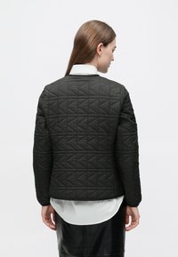 Geacă neagră matlasată cu un model geometric, guler rotund și mâneci lungi, purtată peste o cămașă albă. Textura pare netedă.