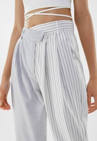 Bílé pruhované kalhoty s kombinací kostkovaného a vertikálního vzoru. Tkanina má hladkou texturu a je opatřena páskem v pase.