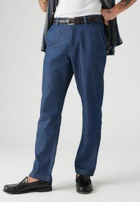 Tummansiniset farkut rentolla leikkauksella, joissa on sivutaskut ja hienovarainen tekstuuri, yhdistetty mustiin nahkakenkiin.