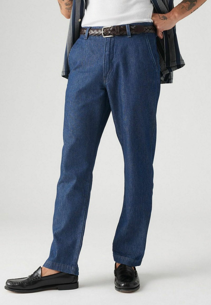 Pantaloni in denim di colore blu scuro con una vestibilità regolare, dotati di tasche laterali, dettagli cuciti, indossati con una cintura intrecciata marrone e scarpe nere.