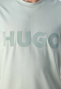 Svetlo zelena majica s kratkimi rokavi z velikimi, poudarjenimi velikimi črkami, ki po širini prsnega dela črkujejo "HUGO" v usklajeni barvi.