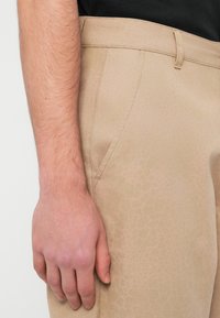 Κλείσιμο της αριστερής παλάμης ενός ατόμου που ακουμπάει πάνω σε μπεζ παντελόνι με διακριτικό εξαγωνικό σχέδιο και ορατή ραφή.