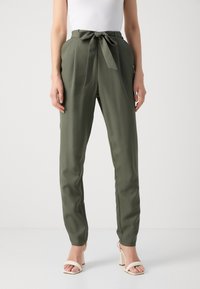 Olivgrüne, hoch taillierte Hose mit einem Stoffgürtel, schmal zulaufenden Beinen und einer glatten Textur. Kombiniert mit hellen Blockabsatzsandalen.