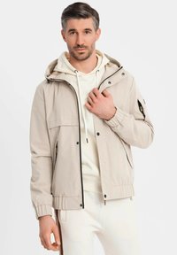 Mann in beiger Kapuzenjacke über einem cremefarbenen Hoodie und passenden Hosen, stehend vor einem schlichten weißen Hintergrund.