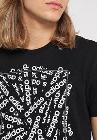 Musta puuvillapaita, jossa on toistuva valkoinen Adidas-logo kierrettyinä ja kuviollisina, täydentää crew-neck-malli.