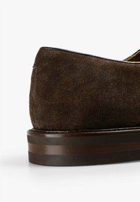 Zapato de ante marrón con una textura suave, diseño estilizado y un tacón de madera oscura que presenta sutiles franjas horizontales de acento.