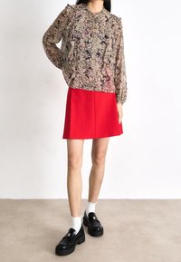 Blusa lunga trasparente con motivo floreale e ruches, abbinata a una gonna A-line rossa. Il modello indossa mocassini neri con calzini bianchi.