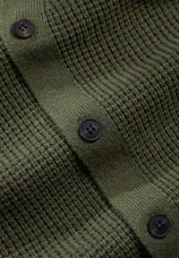 Țesătură tricotată de culoare verde măslinie cu un model texturat. Patru butoane negre sunt vizibile, aranjate vertical de-a lungul marginii.
