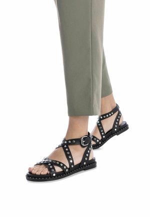 Sandalias de cuero negro con acentos de tachuelas, diseño entrelazado, hebilla ajustable y suela plana, combinadas con pantalones cortos de color verde oliva.