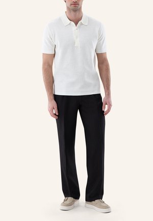 Mann trägt kurzärmeliges weißes Strick-Poloshirt, schwarze gerade Hose und beige Slipper, steht vor schlichtem hellem Hintergrund.