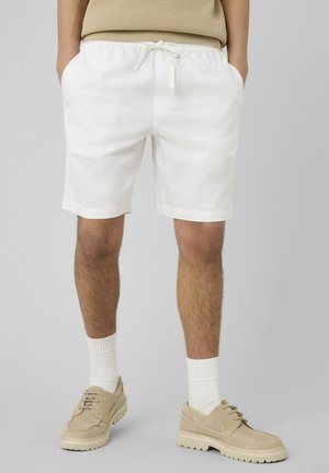 Homme portant un short blanc à cordon, un haut beige en tricot, des chaussettes blanches et des chaussures beiges à lacets, debout devant un fond uni.