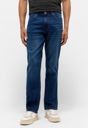 Dark blue denim jeans with a straight-cut design, five pockets, and tonal stitching, paired with beige and white sneakers.