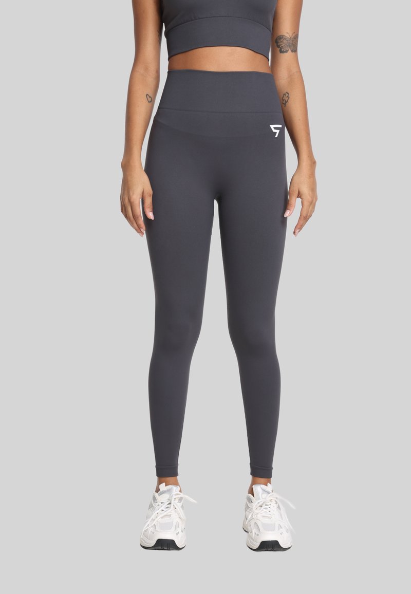 Kobieta ubrana w ciemnoszare legginsy z wysokim stanem oraz dopasowany top, nosząca białe buty sportowe, na jednolitym jasnym tle.