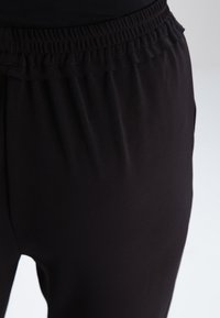 Black elastic-waist pants with a smooth texture. They feature a gathered waistband and a close-fit along the legs. No visible pockets.
